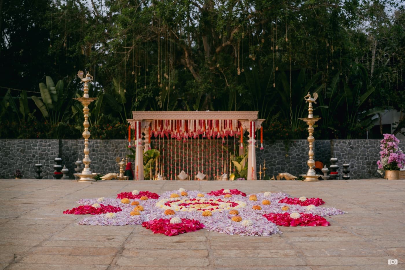 A mandap decorated with marigold and rose petal rangoli, brass lamps, and hanging floral strings