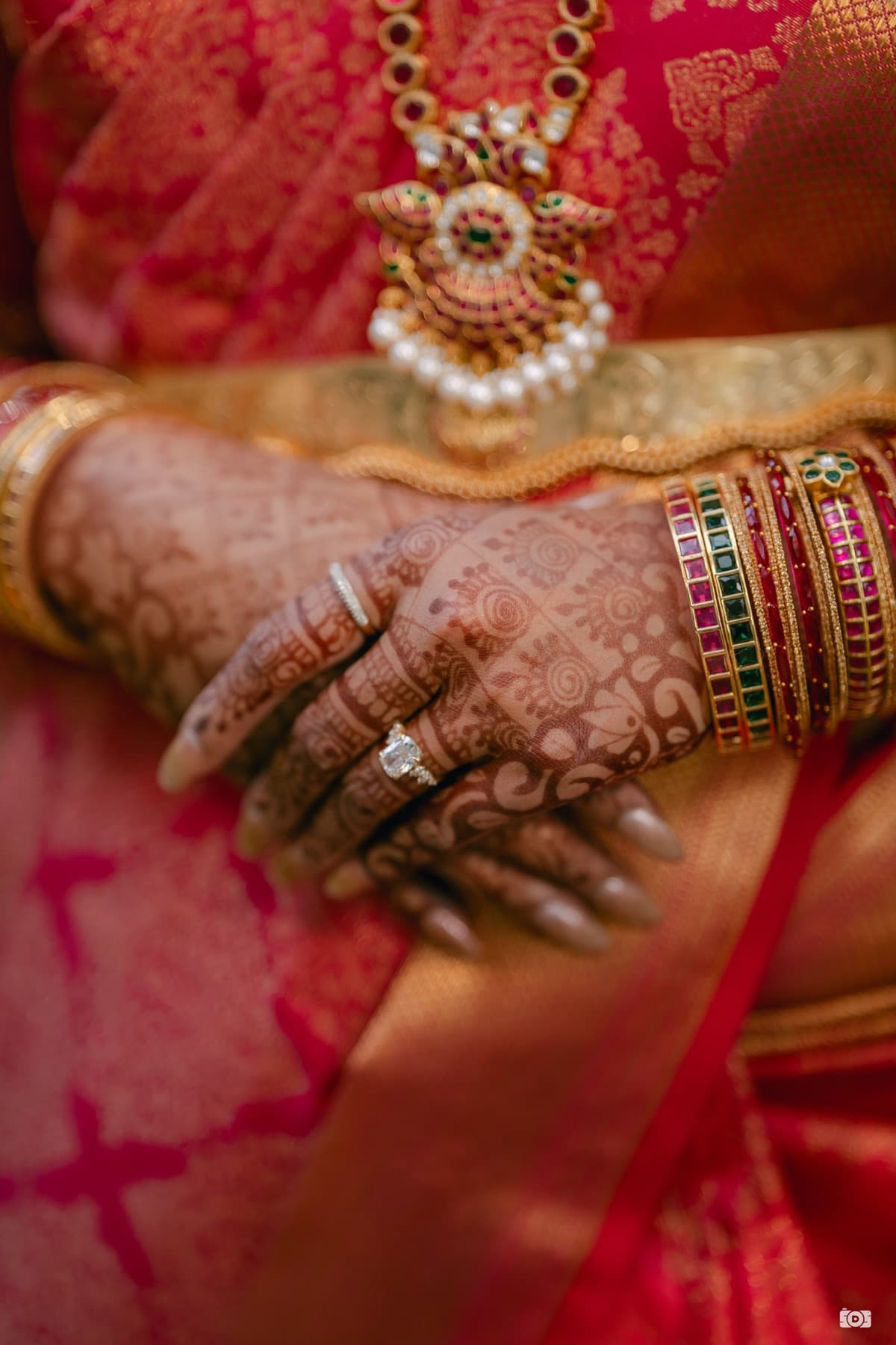 A bride's mehndi-adorned hands with engagement and wedding rings, layered bangles, and a gold necklace pendant